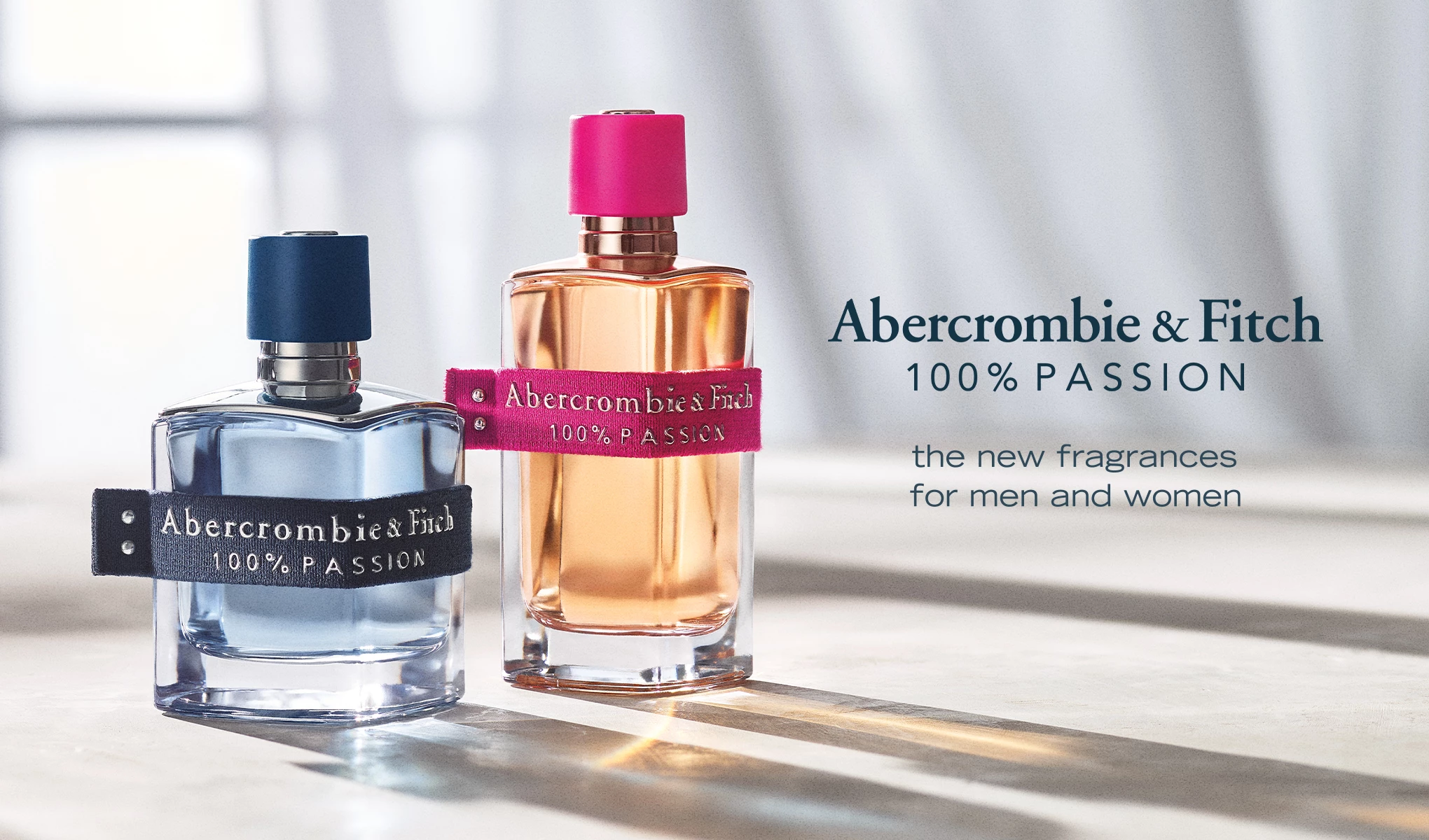 To parfymeflasker fra Abercrombie & Fitch med teksten "100% PASSION". Den ene flasken er blå med en mørkeblå etikett, og den andre er oransje med en rosa etikett.