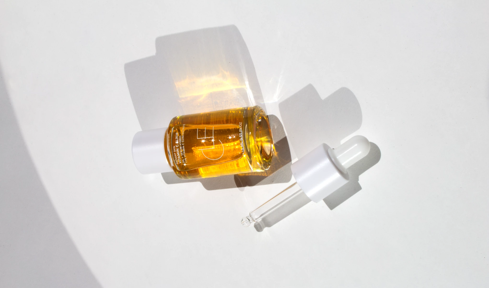 Pieni keltaista nestettä sisältävä pullo, jossa on merkintä ”Vitamin C Elixir”, makaa valkoisella alustalla. Pullossa on valkoinen korkki ja sen vieressä pipetti.