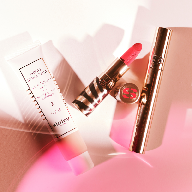 Sisley Phyto Hydra Teint farget dagkrem og rød leppestift på hvit bakgrunn med mykt rosa lys.