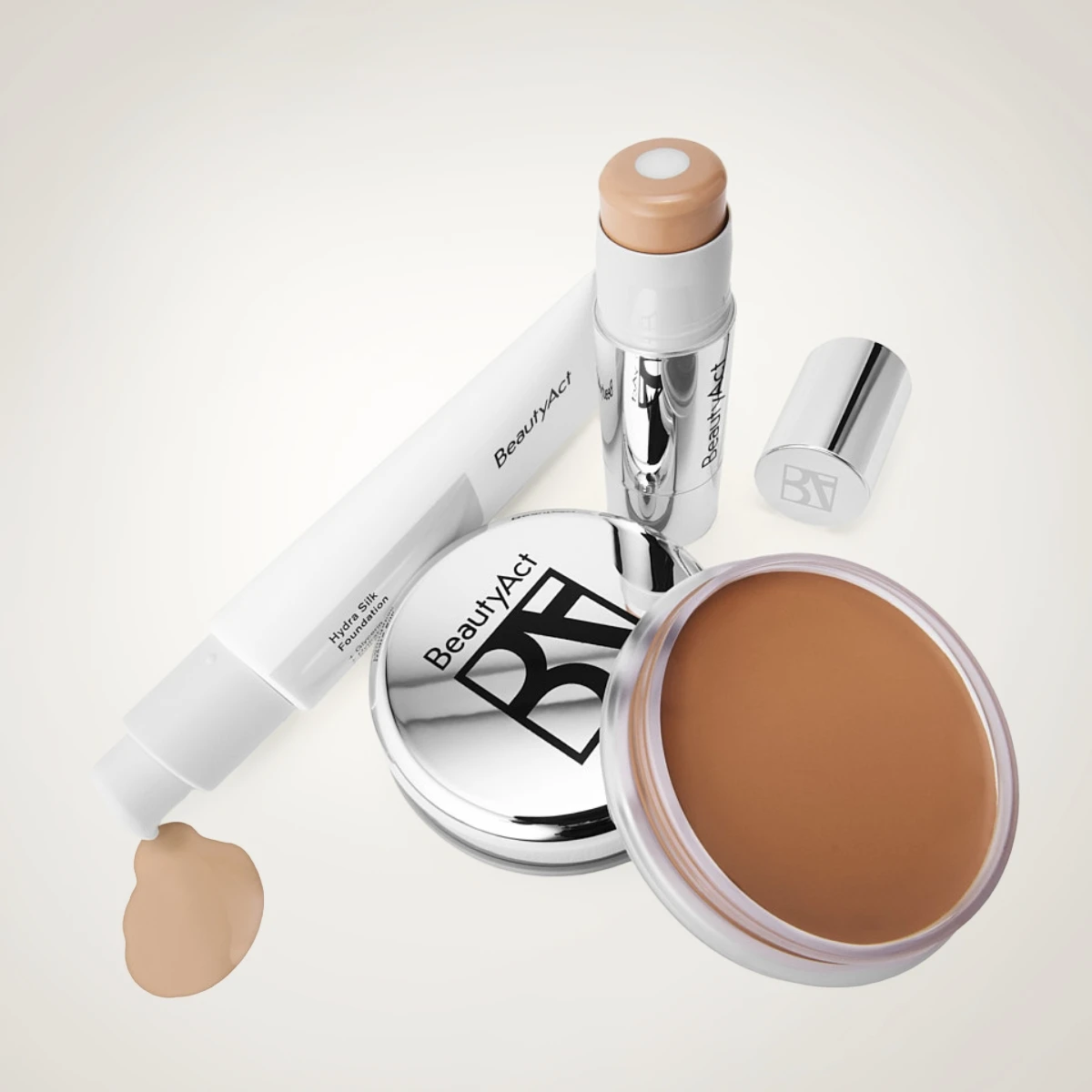 BeautyAct makeup-produkter med foundation och bas: tub märkt “BeautyAct Hydra Silk Foundation”, stickfoundation, kompakt med BA-logotyp och burk med krämig foundation i brun nyans på ljus bakgrund.