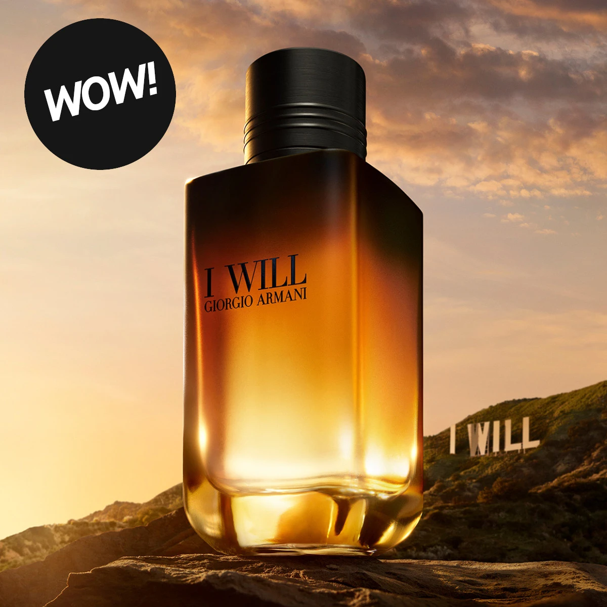 Giorgio Armani I WILL parfyme i en stor ravfarget flaske med svart kork, plassert på en klippe foran et fjellandskap i solnedgang. Teksten «I WILL» vises på fjellet og «WOW!» i en svart sirkel.