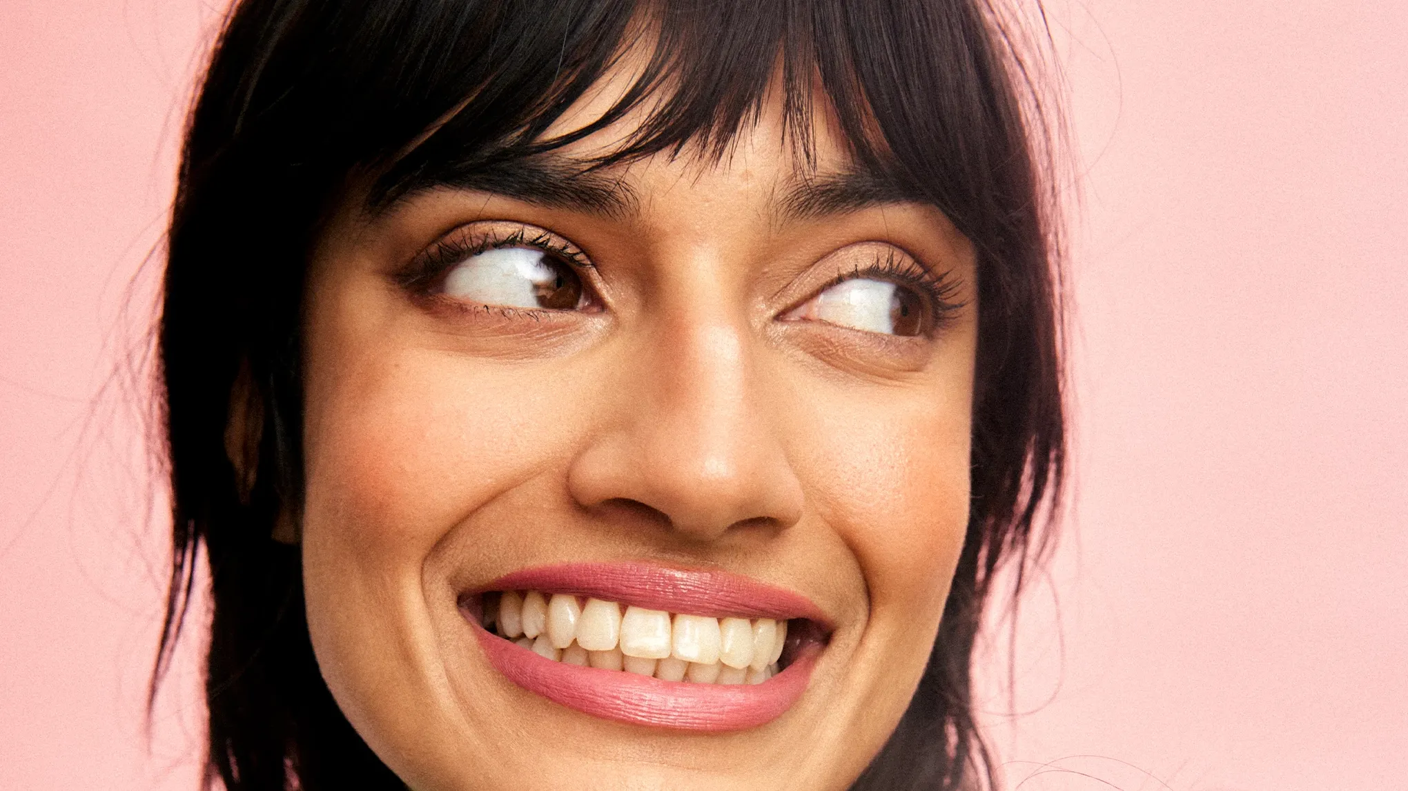 Nærbilde av en smilende person med mørkt hår og rosa leppestift mot en rosa bakgrunn.