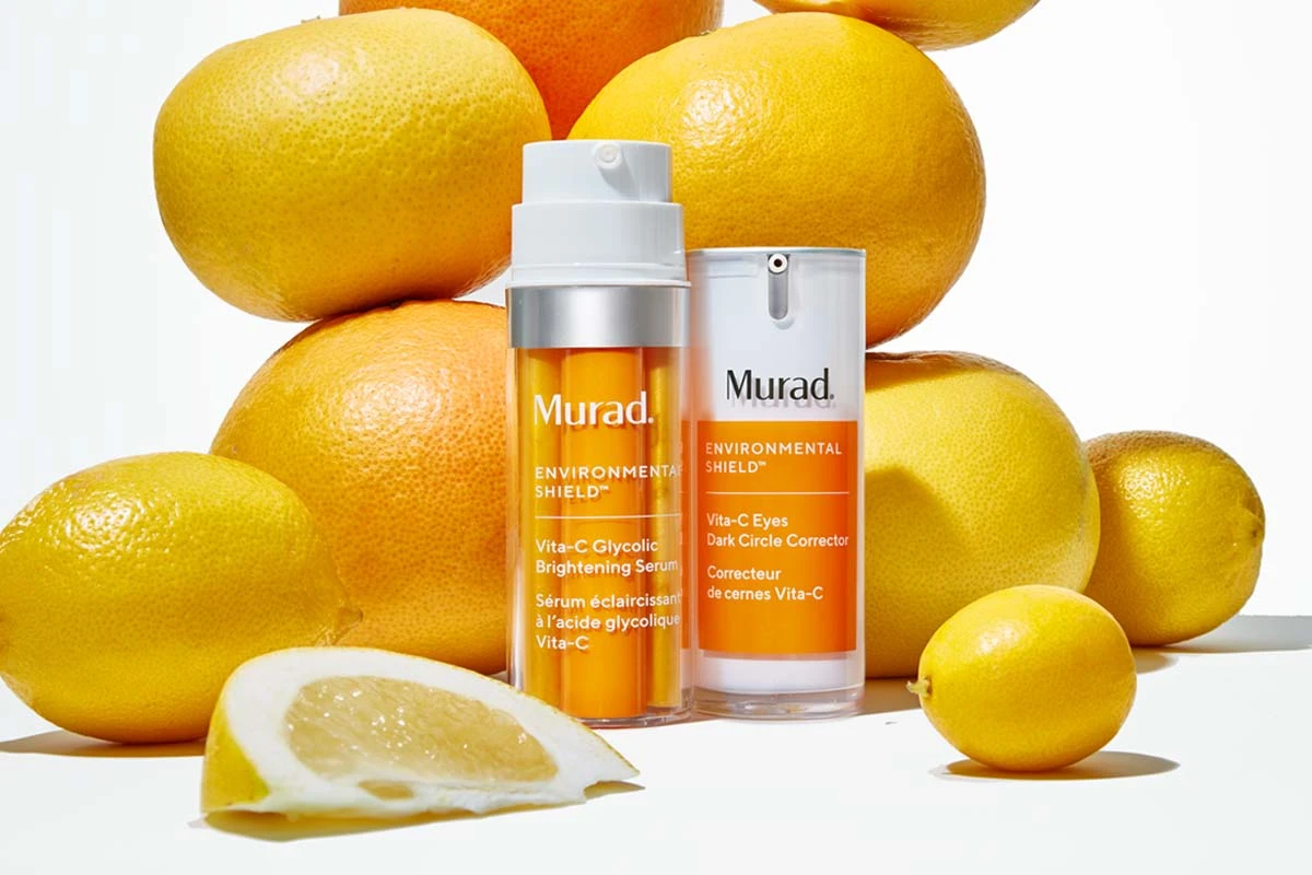 Två produkter från Murad placerade framför en hög med citrusfrukter, inklusive citroner och apelsiner. Produkterna har orange etiketter och är omgivna av frukterna, vilket framhäver deras C-vitamin innehåll och hudvårdande egenskaper.