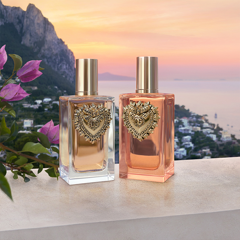 Två parfymer från Dolce & Gabbana, placerade på en stenliknande yta. Flaskorna har ett dekorativt hjärtemblem och är fyllda med gyllene och rosa vätska. Bakgrunden visar en vacker kuststad vid solnedgången, med havet och berg i fjärran. Blommor med rosa kronblad syns till vänster i bilden.