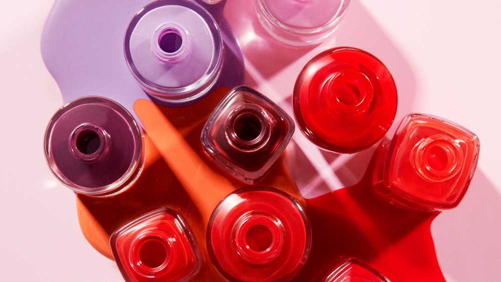 Useita avonaisia Atelier Rouge -kynsilakkapulloja ylhäältä kuvattuna, väriläiskiä punaisena, oranssina ja violettina vaaleanpunaisella taustalla.