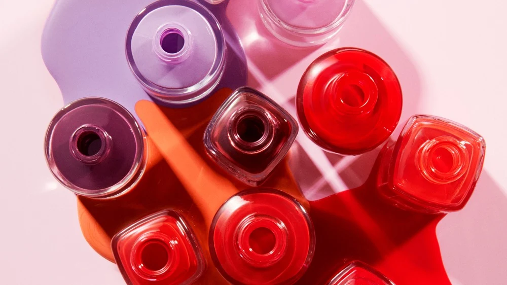 Useita avonaisia Atelier Rouge -kynsilakkapulloja ylhäältä kuvattuna, väriläiskiä punaisena, oranssina ja violettina vaaleanpunaisella taustalla.
