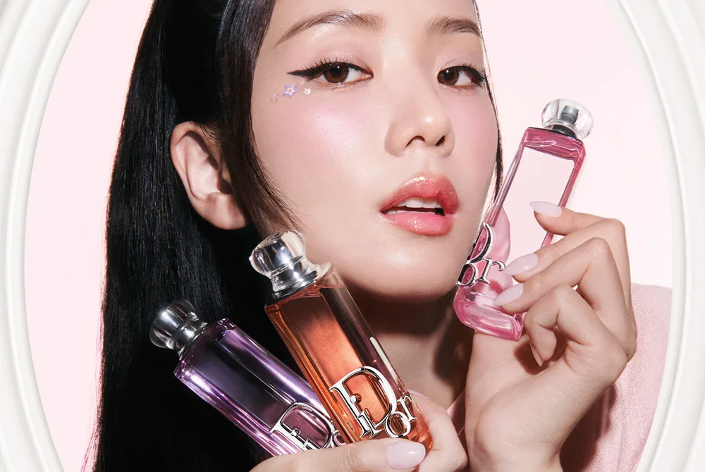 Kvinna håller tre Dior Addict Lip Glow Oil i olika nyanser framför ansiktet mot en ljusrosa bakgrund.