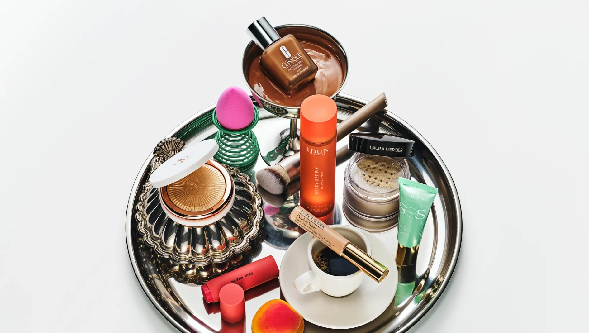 Makeup och hudvård arrangerade på en silverfärgad bricka tillsammans med en kaffekopp och frukt. Produkterna inkluderar Clinique foundation, IDUN Minerals Ready Set Fix Setting Spray, Laura Mercier puder, Estée Lauder Double Wear concealer, Clarins primer och Milani Jord läpprodukt.
