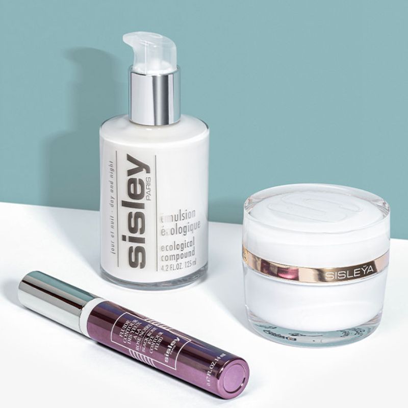 Tre hudvårdsprodukter från Sisley placerade på en vit yta med en blå bakgrund. En flaska med pump, en burk samt en tub. Produkterna har förpackningar i vitt, silver och guld.