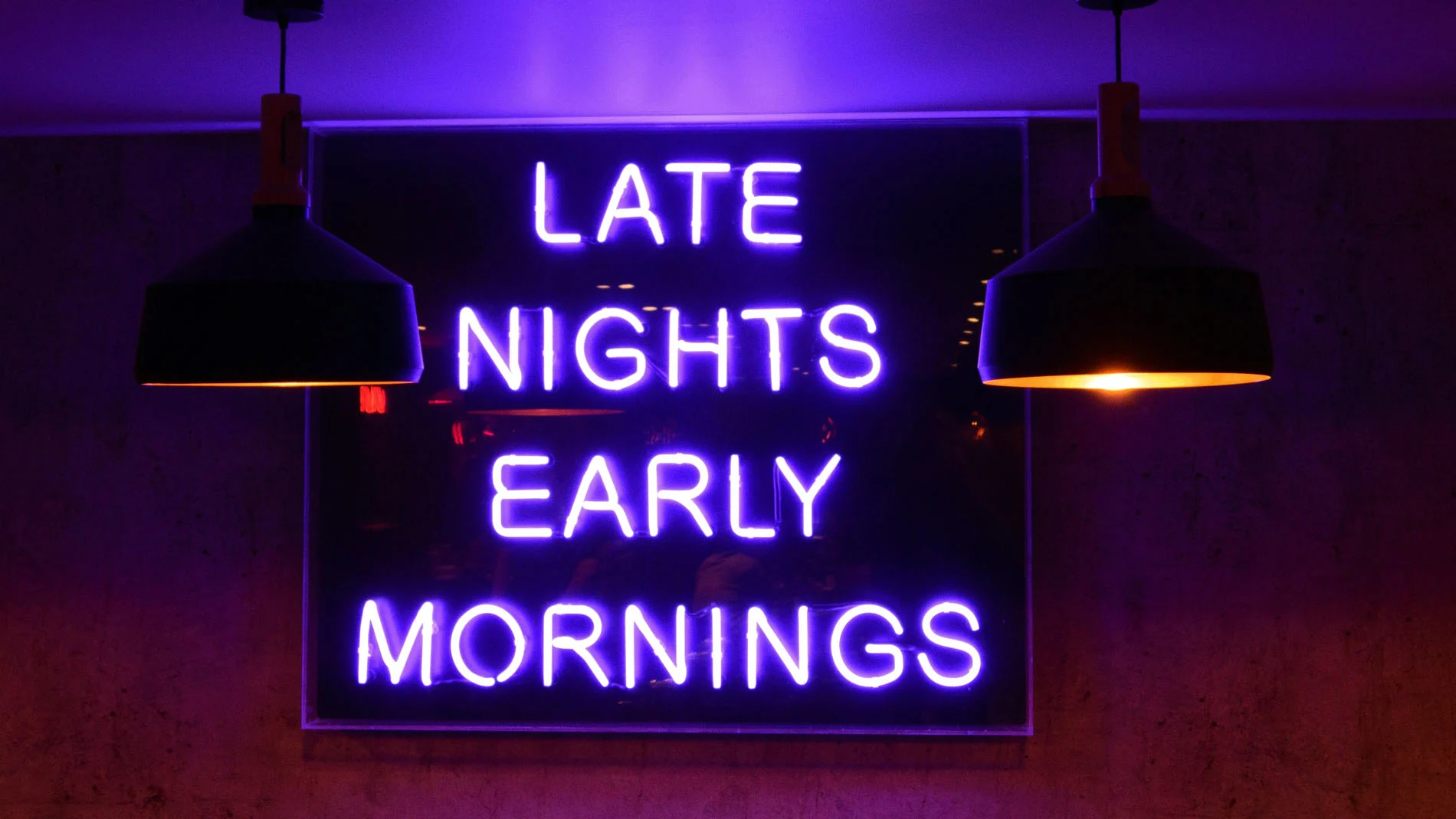 Neonljusskylt med texten "Late Nights Early Mornings" i ett mörkt rum, upplyst av två hängande lampor