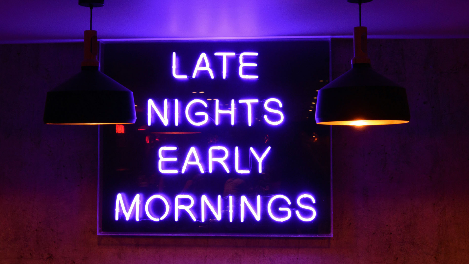 Neonvalokyltti, jossa lukee "Late Nights Early Mornings" pimeässä huoneessa, valaistuna kahdella roikkuvalla lampulla