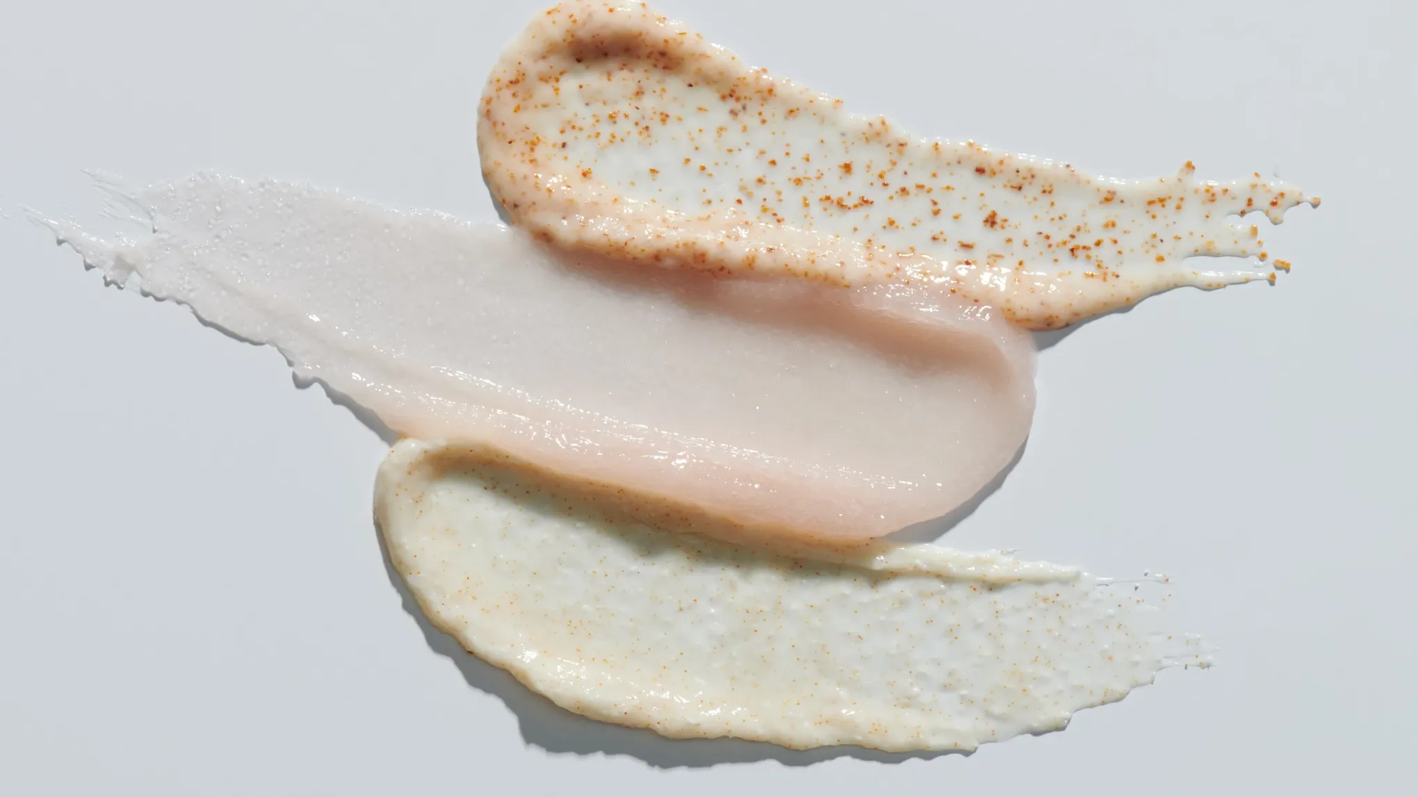 Tre olika texturer av hudvårdsprodukter, inklusive krämiga och geléliknande konsistenser med exfolierande korn, utsmetade på en vit bakgrund.