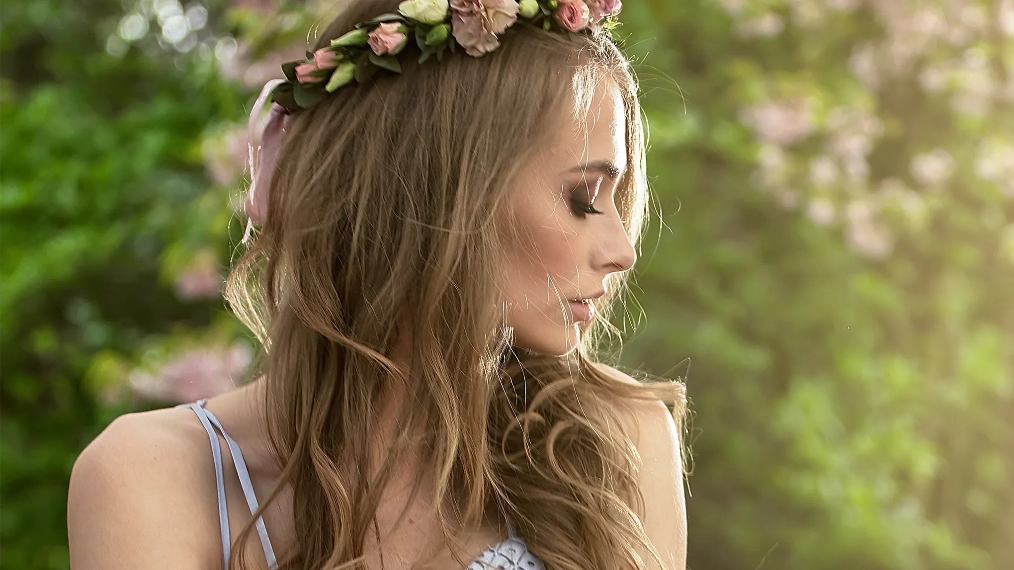 En kvinna med långt hår och en blomkrans på huvudet, i profil framför gröna träd.