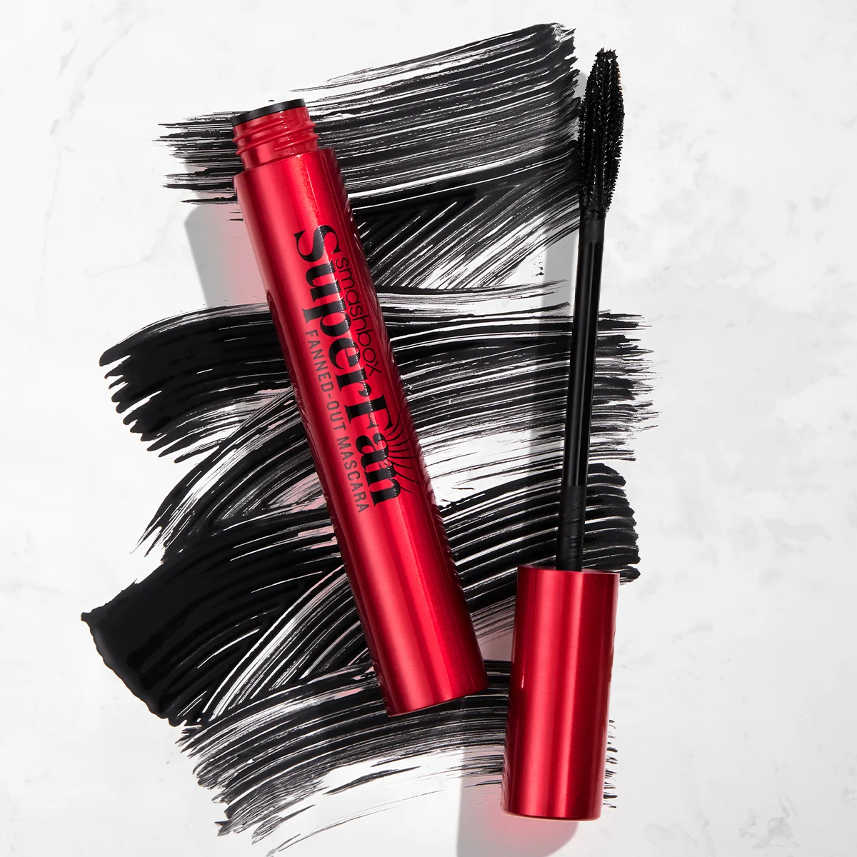 Smashbox Superfan Fanned-Out Mascara i en röd tub, med mascaraborsten bredvid och svarta mascarastråk i bakgrunden.