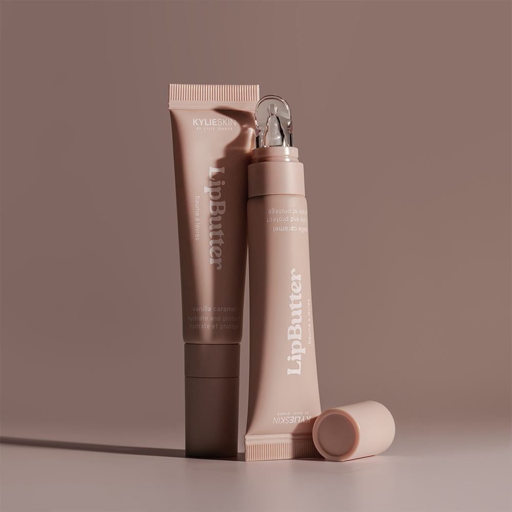 Kaksi Kylieskin LipButter-tuubia seisovat tyylikkäästi ruskealla taustalla. Tuubit ovat vaaleanruskeita ja sisältävät vanilja-karamellin makuista huulivoidetta.