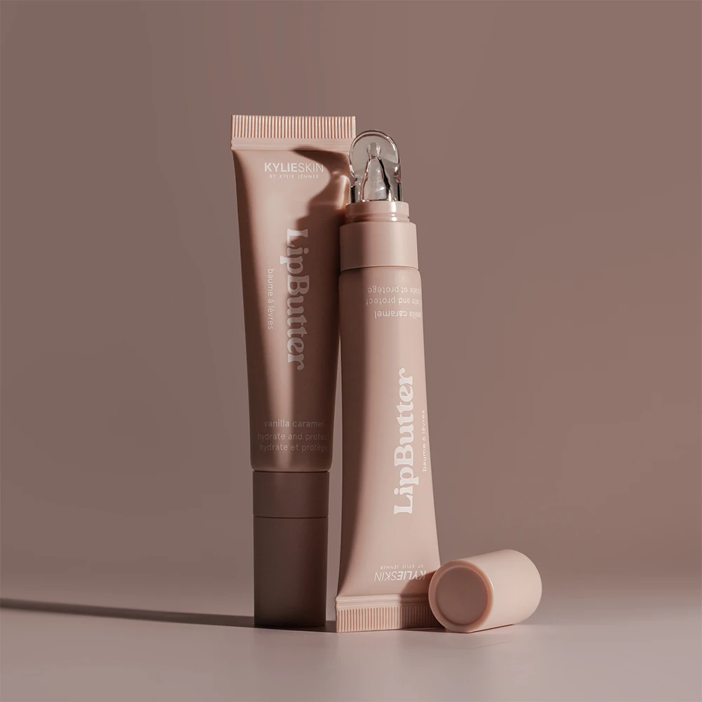 Kaksi Kylieskin LipButter-tuubia seisovat tyylikkäästi ruskealla taustalla. Tuubit ovat vaaleanruskeita ja sisältävät vanilja-karamellin makuista huulivoidetta.