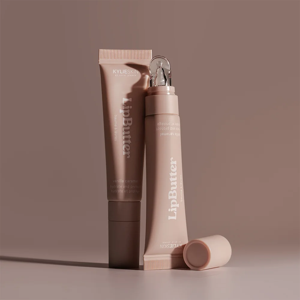 Kaksi Kylieskin LipButter-tuubia seisovat tyylikkäästi ruskealla taustalla. Tuubit ovat vaaleanruskeita ja sisältävät vanilja-karamellin makuista huulivoidetta.
