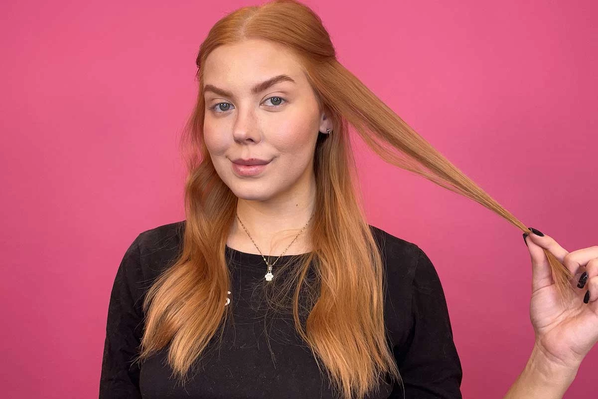 En person med långt, rött hår står framför en rosa bakgrund och har på sig en svart tröja med texten "KICKS". Personen håller i en hårslinga.