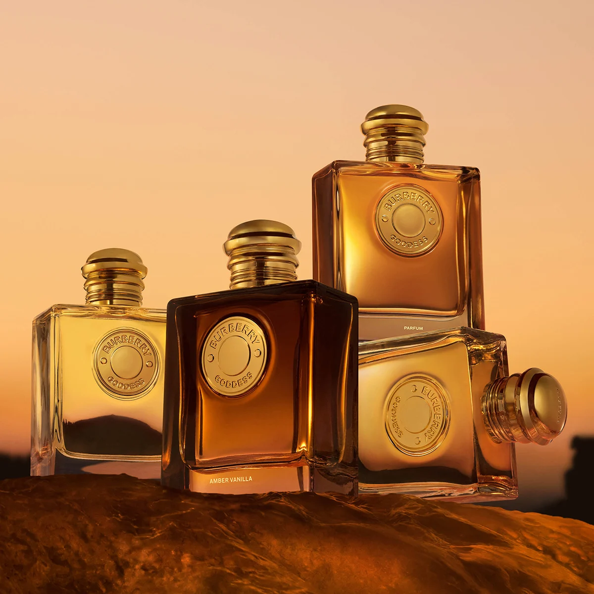 Neljä Burberry Goddess -hajuvesipulloa suorakulmaisissa lasipulloissa, joissa on kullanvärisiä yksityiskohtia, lämpimän oranssia taustaa vasten. Pulloissa näkyvät tekstit ”BURBERRY GODDESS”, ”PARFUM” ja ”AMBER VANILLA”.