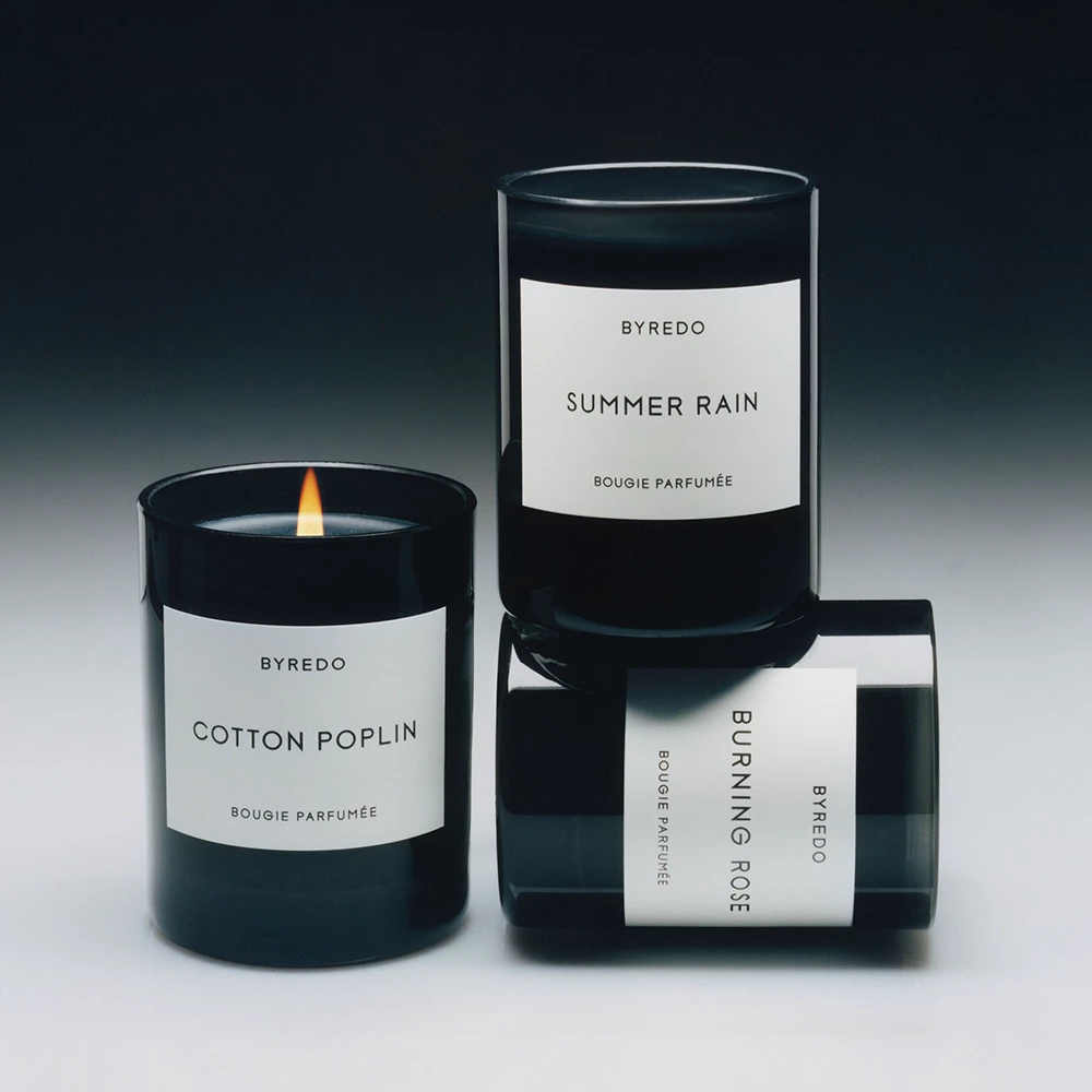 Tre doftljus från Byredo, arrangerade på en vit yta med en mörk bakgrund. Ljusen har svarta glasbehållare med vita etiketter och namn som "Cotton Poplin", "Summer Rain" och "Burning Rose". Ett av ljusen är tänt.