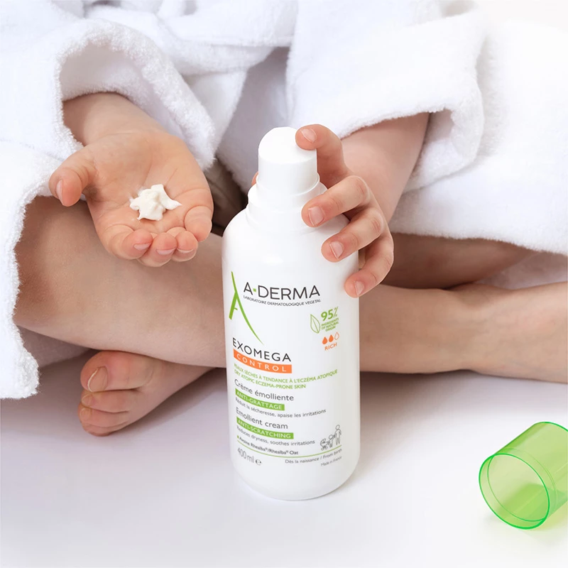 Barn som sitter iklädd en vit badrock och håller en vit flaska med hudkräm från märket A-Derma. Flaskan har texten "Exomega Control Emollient Cream". Barnet har en liten mängd kräm i sin öppna hand. Ett grönt lock ligger bredvid barnet på den vita ytan. Bakgrunden är ljus och enkel, vilket framhäver produkten och barnets händer.