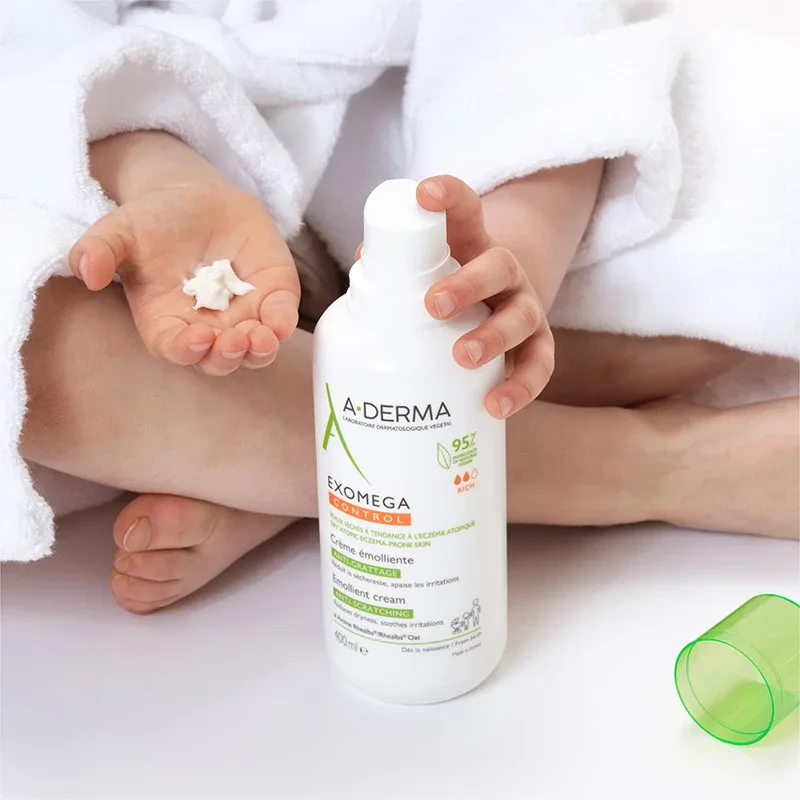 Barn som sitter iklädd en vit badrock och håller en vit flaska med hudkräm från märket A-Derma. Flaskan har texten "Exomega Control Emollient Cream". Barnet har en liten mängd kräm i sin öppna hand. Ett grönt lock ligger bredvid barnet på den vita ytan. Bakgrunden är ljus och enkel, vilket framhäver produkten och barnets händer.