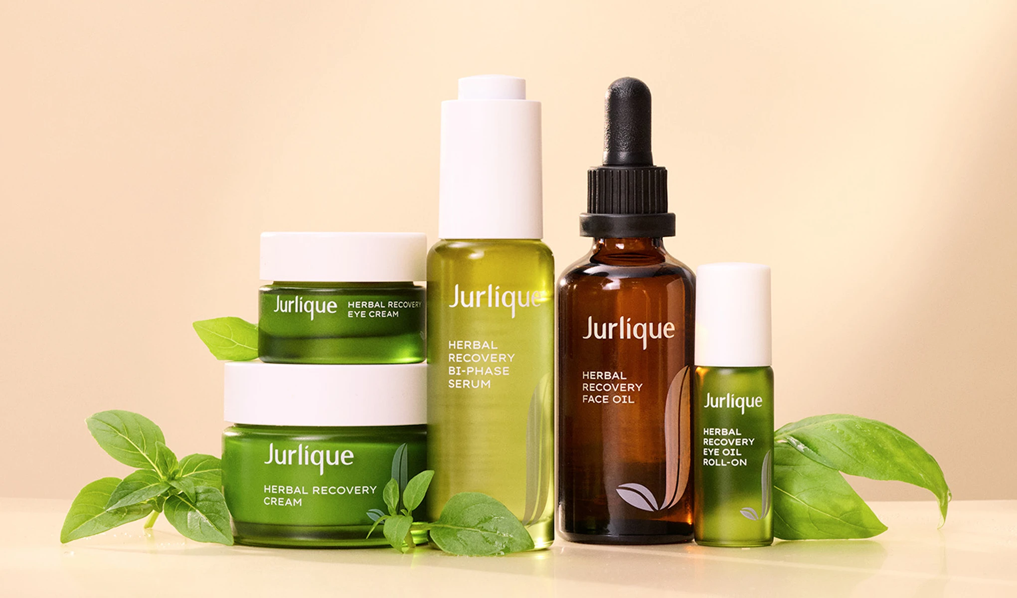 Jurlique-tuotteet, mukaan lukien Herbal Recovery Eye Cream, Herbal Recovery Cream, Herbal Recovery Bi-Phase Serum, Herbal Recovery Face Oil ja Herbal Recovery Eye Oil Roll-On, asetettuna vaalealle taustalle. Tuotteiden ympärillä on vihreitä lehtiä.