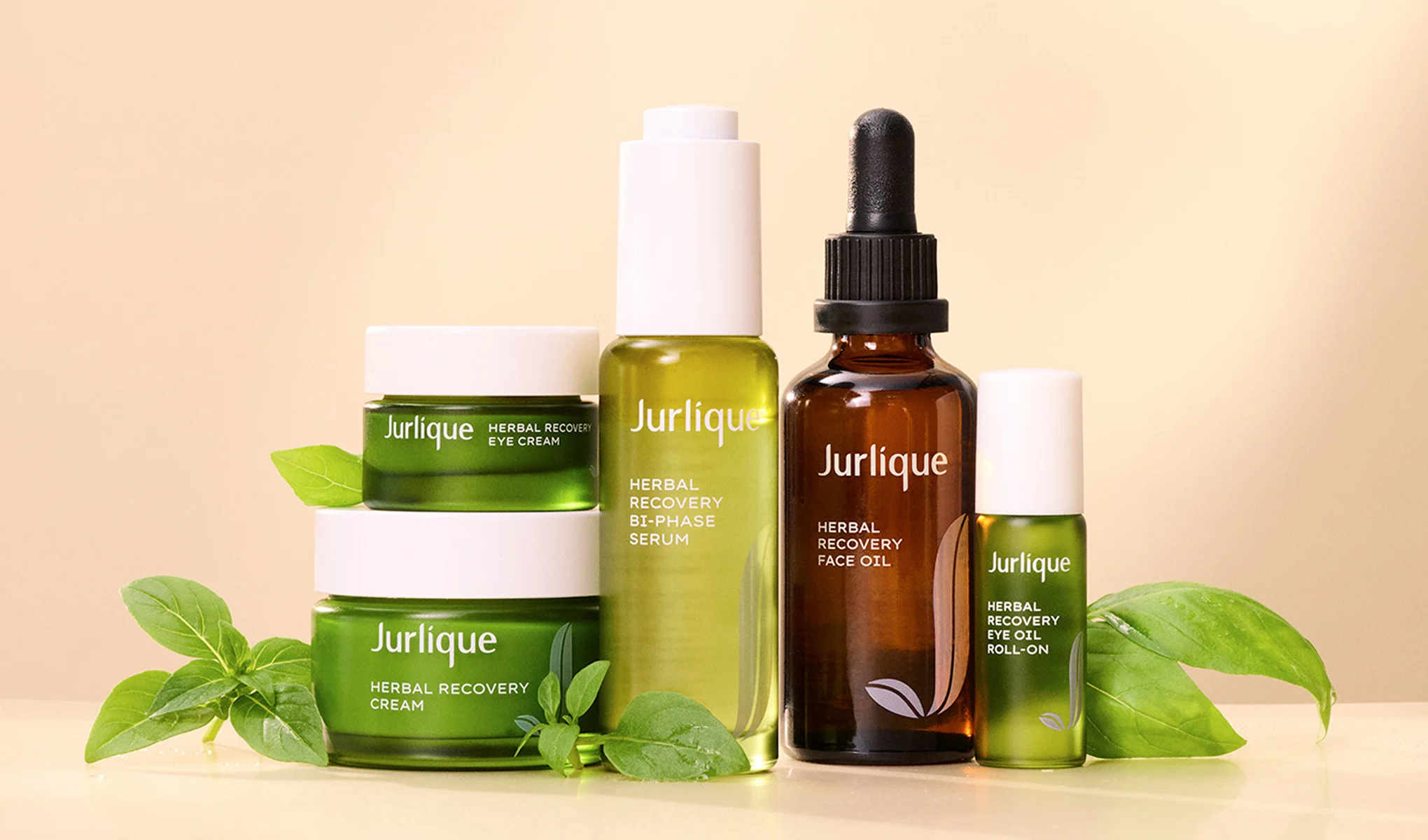 Jurlique-tuotteet, mukaan lukien Herbal Recovery Eye Cream, Herbal Recovery Cream, Herbal Recovery Bi-Phase Serum, Herbal Recovery Face Oil ja Herbal Recovery Eye Oil Roll-On, asetettuna vaalealle taustalle. Tuotteiden ympärillä on vihreitä lehtiä.