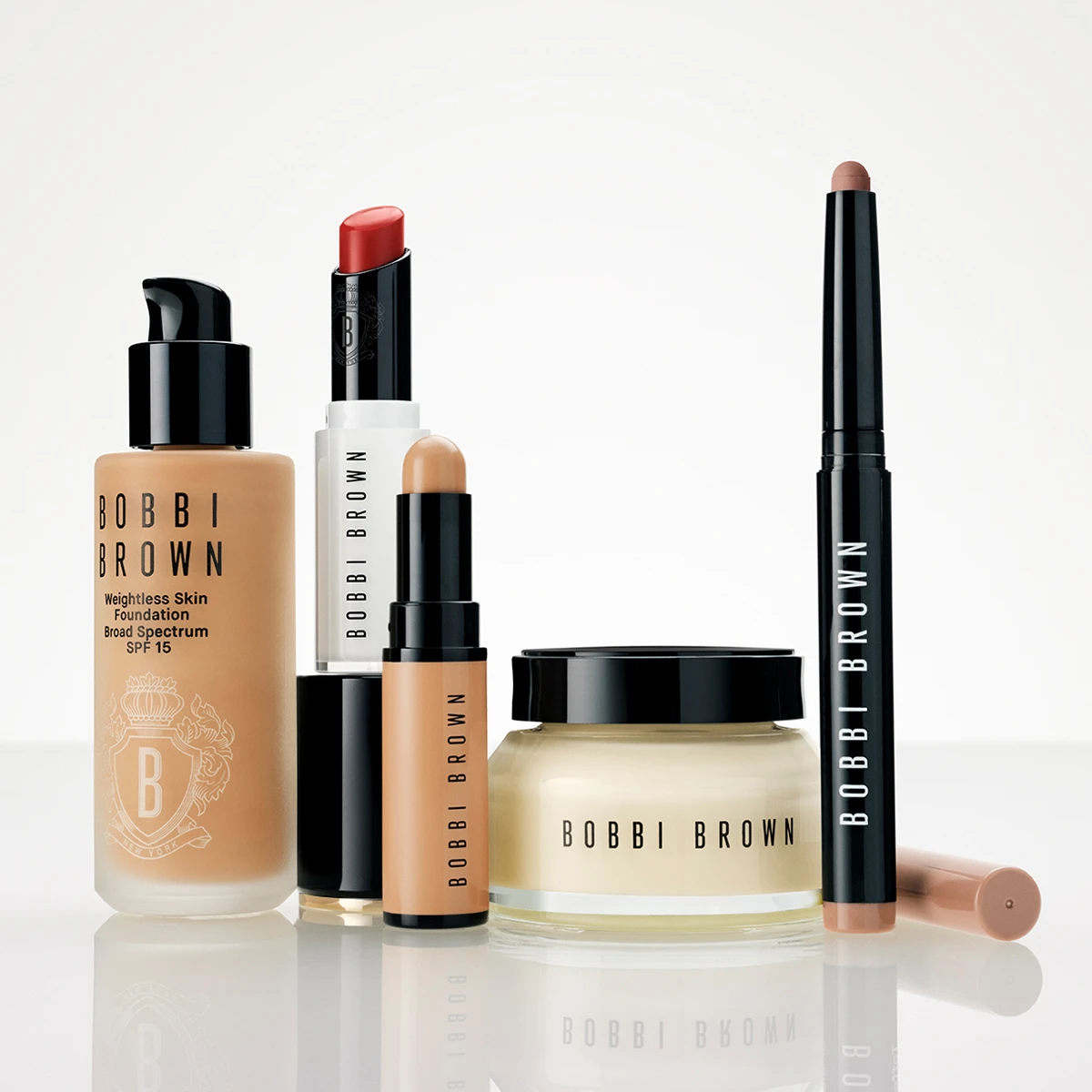 Flera Bobbi Brown-sminkprodukter står tillsammans mot en ljus bakgrund: Weightless Skin Foundation Broad Spectrum SPF 15, en läppstiftshylsa, ett stick-format smink, en ansiktskräm i burk och en sminkpenna. Synlig text: "BOBBI BROWN", "Weightless Skin Foundation", "Broad Spectrum SPF 15", "SPF 15" och "NEW YORK".