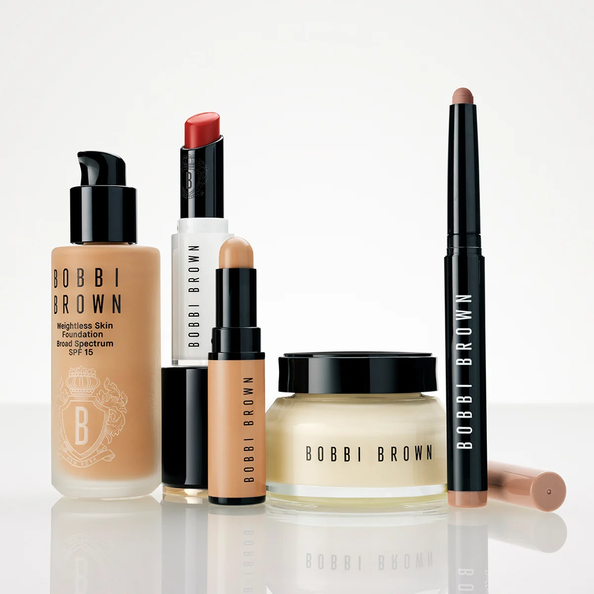 Flera Bobbi Brown-sminkprodukter står tillsammans mot en ljus bakgrund: Weightless Skin Foundation Broad Spectrum SPF 15, en läppstiftshylsa, ett stick-format smink, en ansiktskräm i burk och en sminkpenna. Synlig text: "BOBBI BROWN", "Weightless Skin Foundation", "Broad Spectrum SPF 15", "SPF 15" och "NEW YORK".