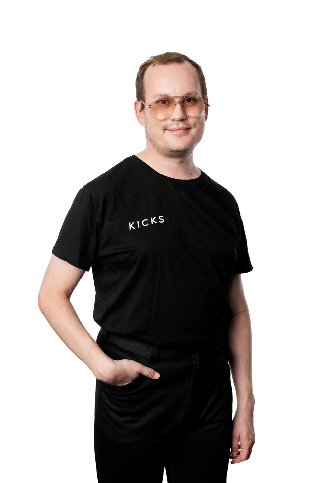 Mies, jolla on silmälasit ja musta T-paita, jossa on KICKS-logo, käsi taskussa, valkoista taustaa vasten.