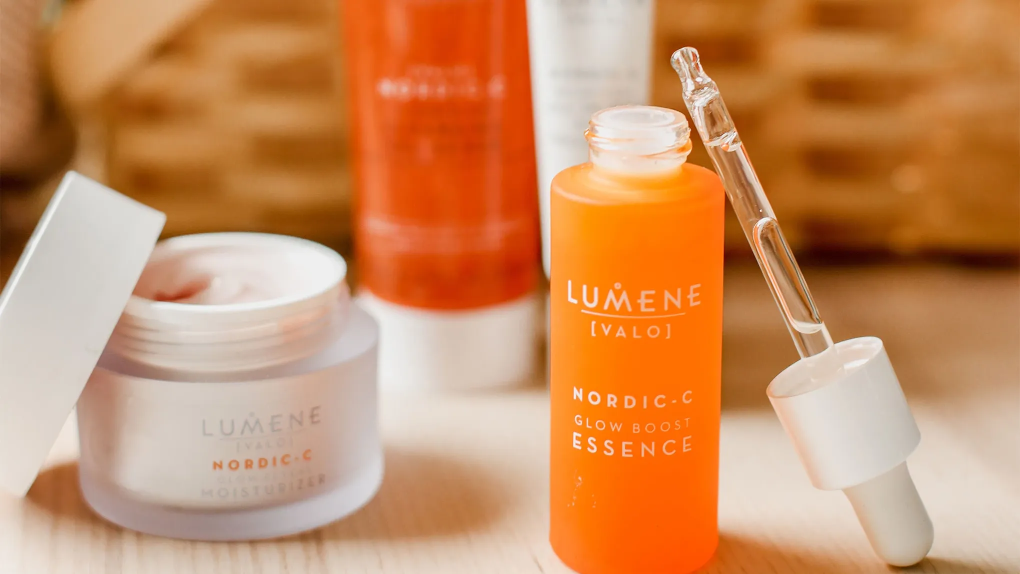 Lähikuva Lumene Nordic-C Glow Boost Essence -tuotteesta ja Nordic-C Glow Reveal Moisturizer -voiteesta. Oranssi pullo pipetillä sisältää essenssin, kun taas valkoinen purkki sisältää kosteusvoiteen. Taustalla näkyy lisää Lumene-tuotteita.