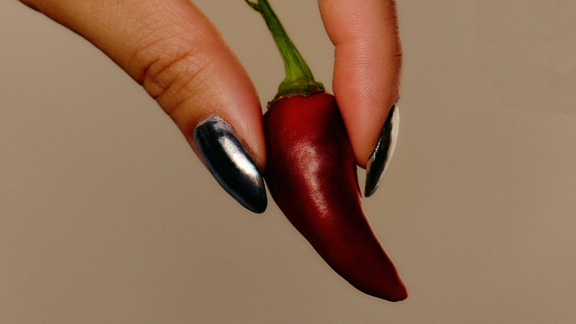 Nærbilde av en hånd med mørk metallic manikyr som holder en rød chilipepper mot en beige bakgrunn. Bildet har en minimalistisk og editorial stil med fokus på negler og sterke fargekontraster.