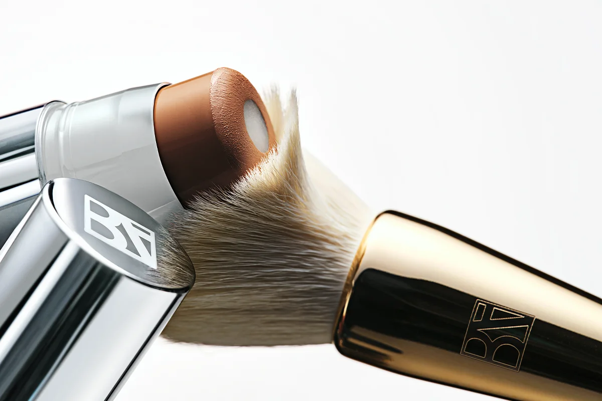 Lähikuva BeautyAct-meikkituotteista, ruskeasta meikkivoidepuikosta ja tiheästä meikkisiveltimestä valkoista taustaa vasten. BeautyAct-logo näkyy siveltimen hopean- ja kullanvärisissä yksityiskohdissa.