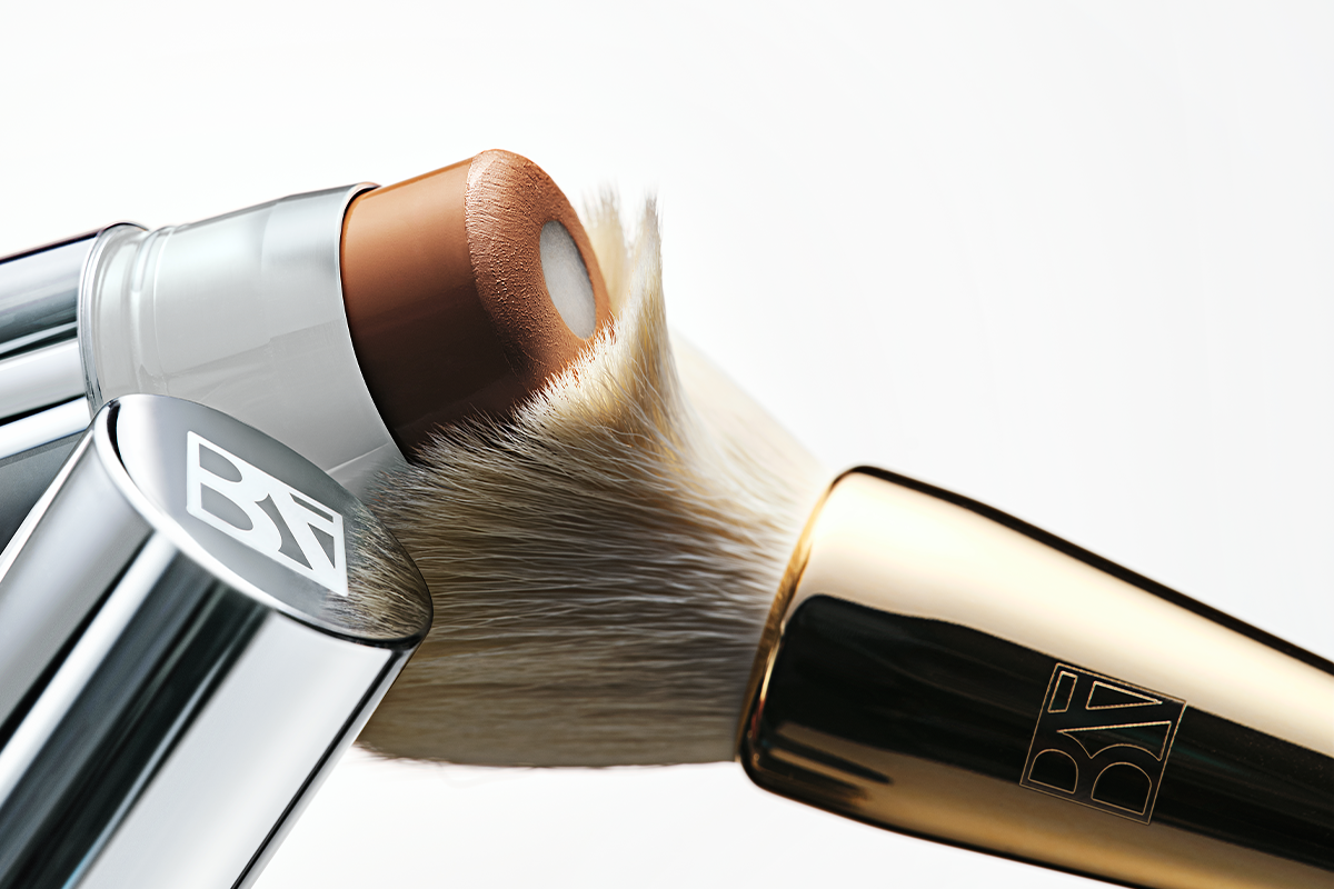 Närbild på BeautyAct makeup med ett brunt foundationstick och en tät sminkborste mot vit bakgrund. BeautyAct-logotypen syns på borstens silver- och guldfärgade detaljer.