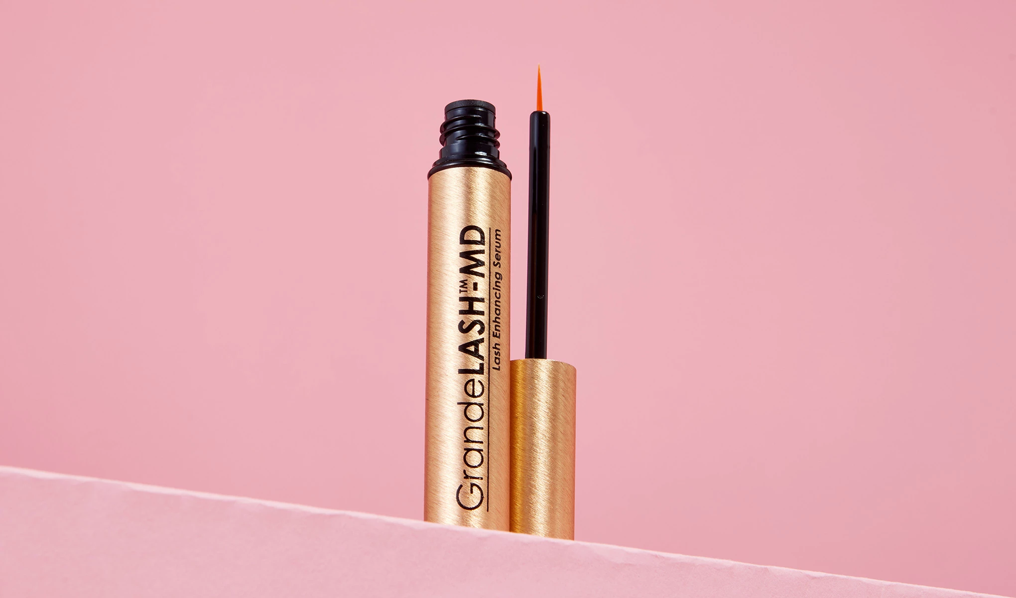 En åpen gullfarget tube med GrandeLASH-MD vippeserum, med en tynn penselapplikator, plassert mot en myk rosa bakgrunn.