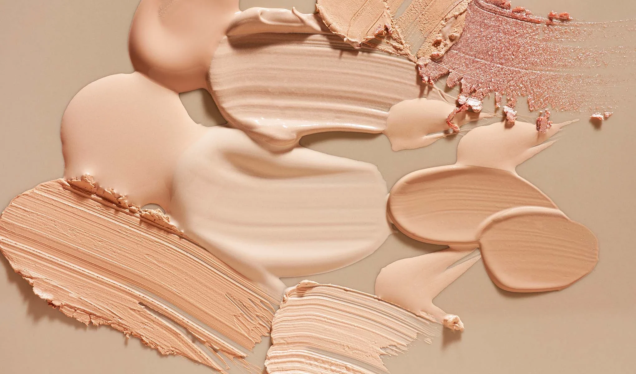 Nærbilde av forskjellige nyanser av foundation og sminke smurt ut på en beige overflate.