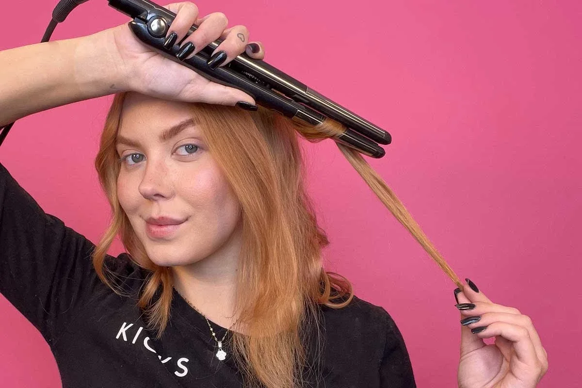 En person med langt, rødt hår står foran en rosa bakgrunn og har på seg en svart genser med teksten "KICKS". Personen bruker en rettetang for å style håret.