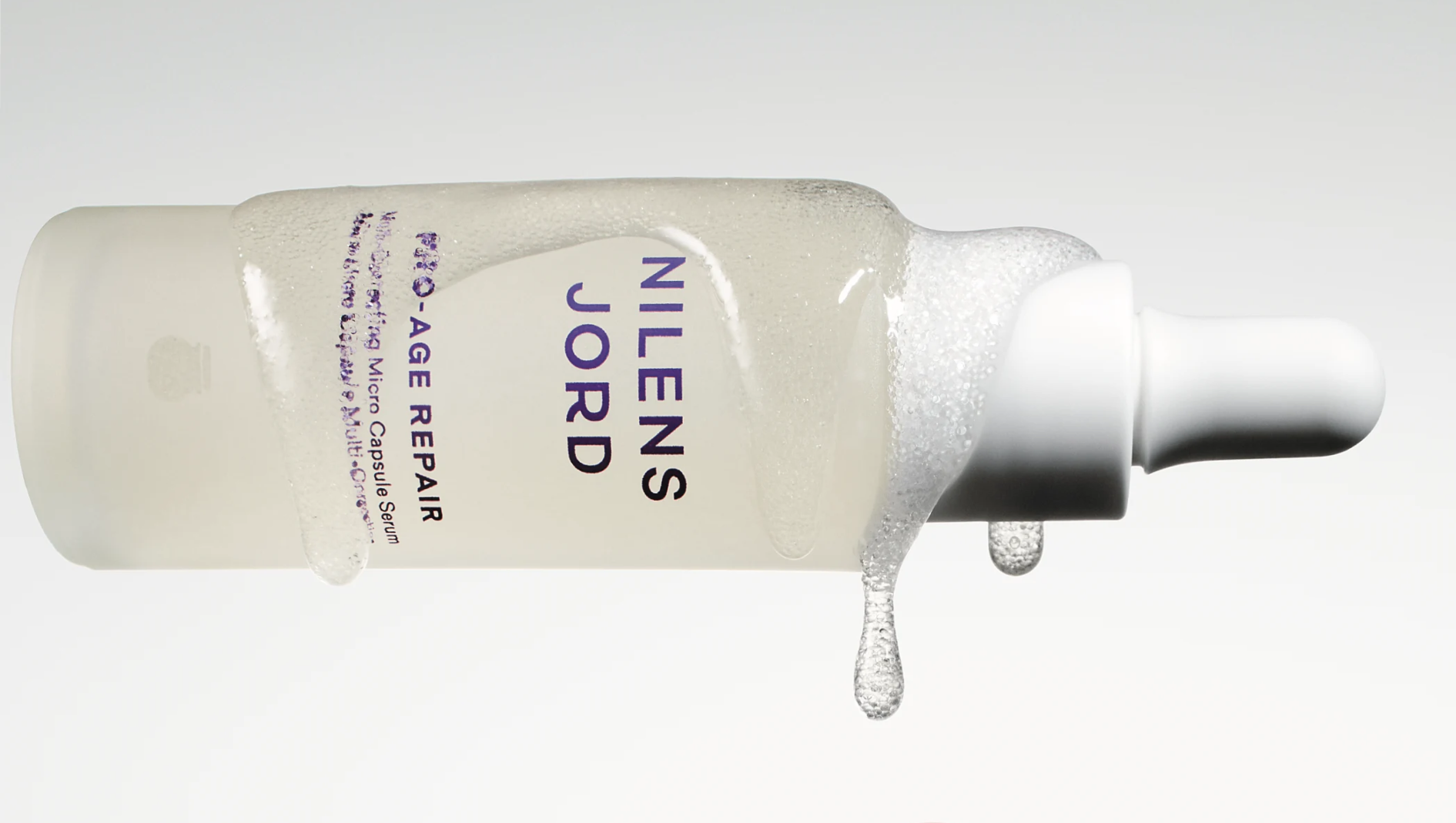 NILENS JORD BIO-AGE REPAIR Micro-Optimising Micro Capsule Serum-flaske, liggende horisontalt med produkt som renner og drypper.