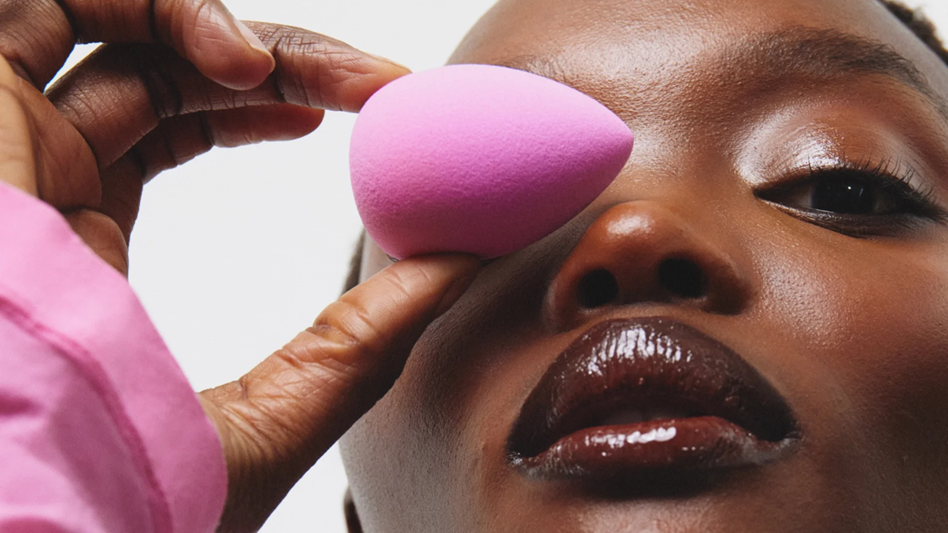 En person i rosa polotröja håller en rosa makeupsvamp mot området kring ögat. Närbild med glansig makeup på läppar och ögon.