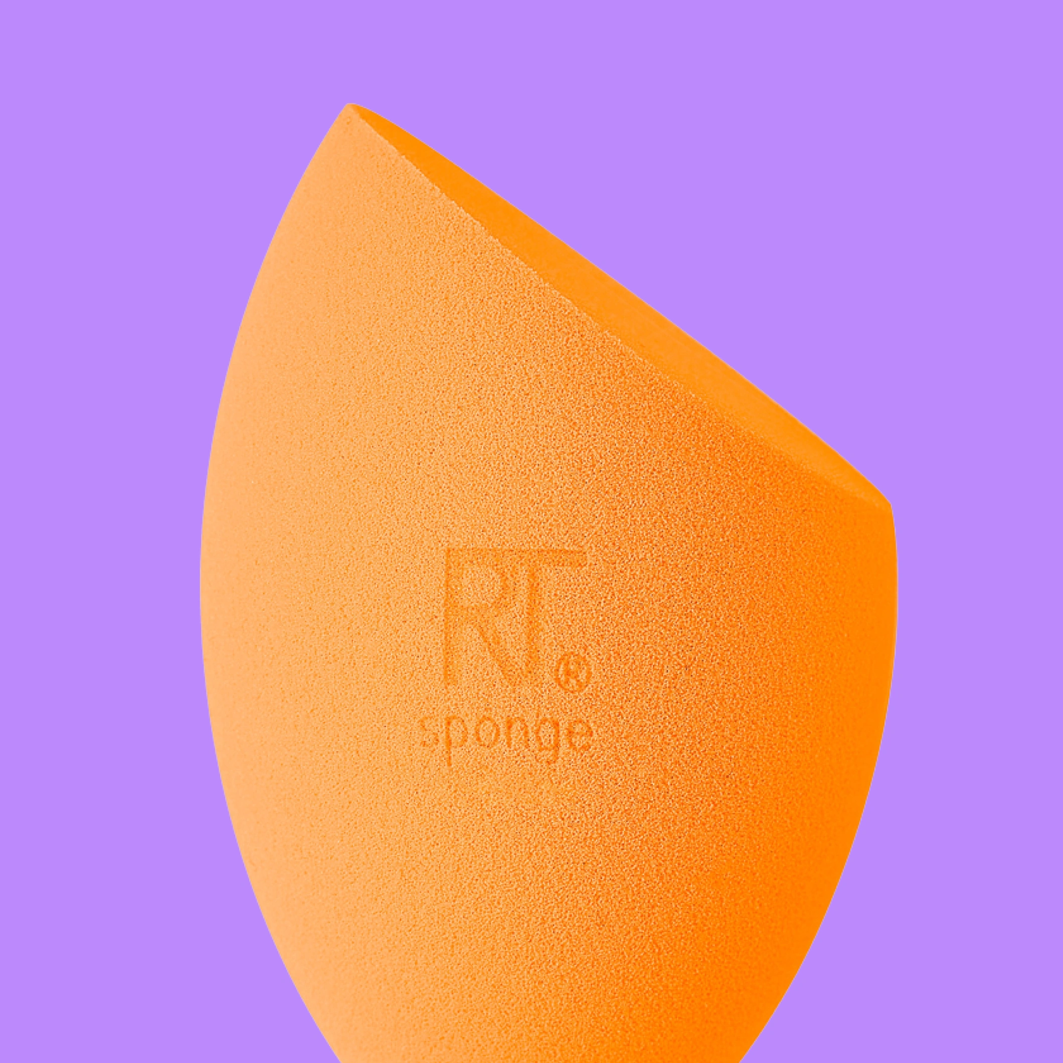 Lähikuva oranssista meikkisienestä ja tiheästä meikkisiveltimestä kuparinvärisellä varrella violettia taustaa vasten. Välineet esitetty erillään ja yksityiskohtaisesti.