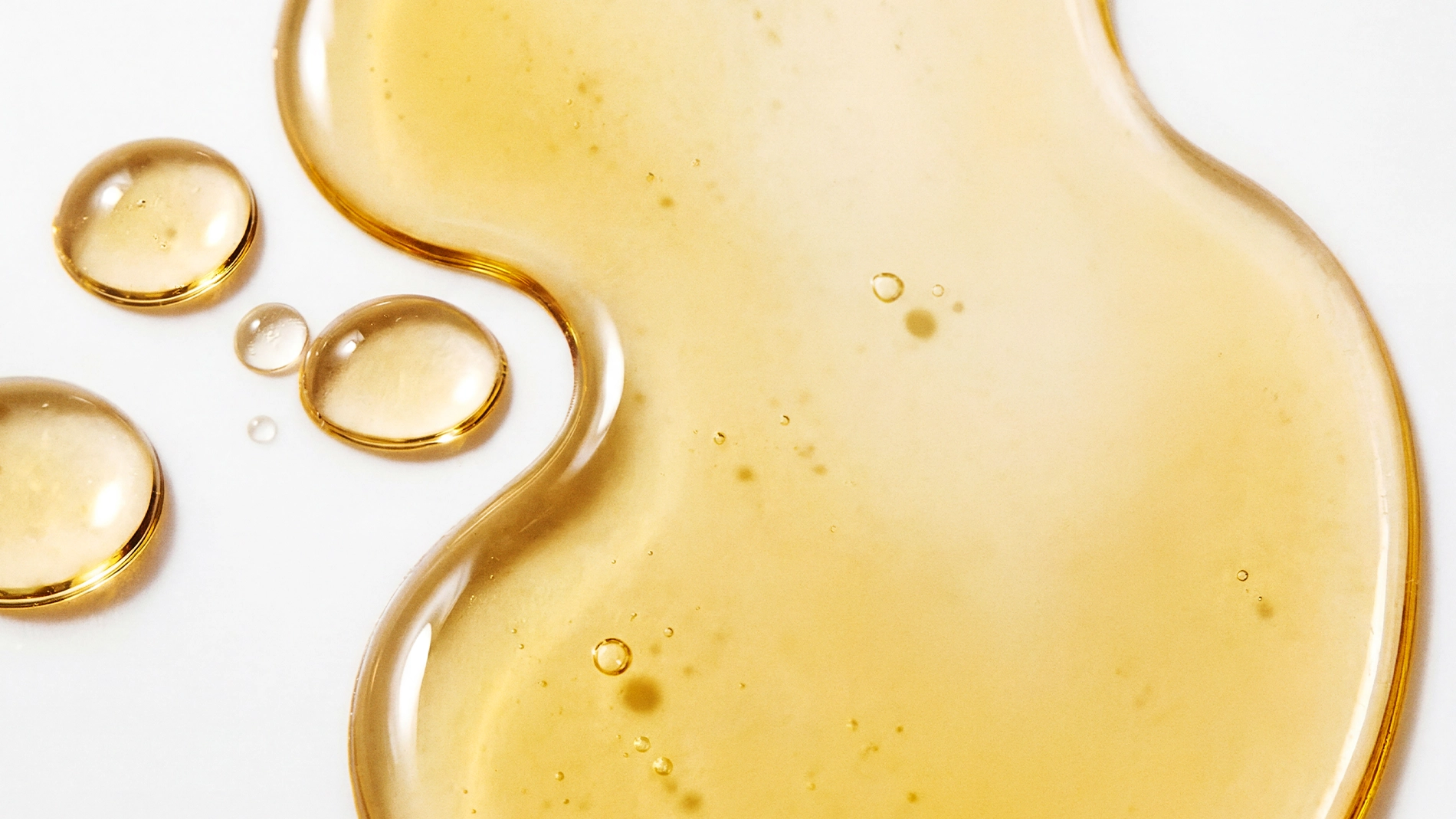 Närbild av gyllene ansiktsolja eller serum i flytande form med droppar och mjuka former på en ljus bakgrund, med fokus på hudvård och återfuktande serum.