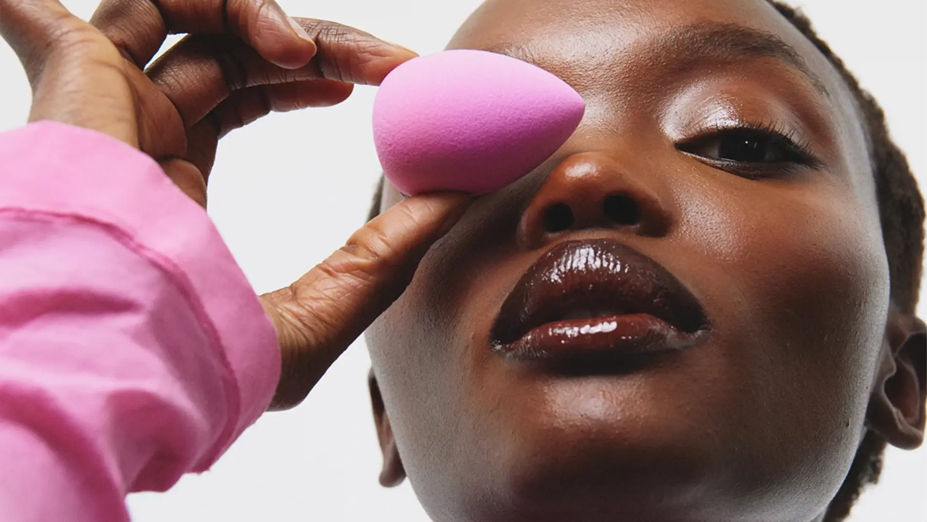 Närbild av en person i rosa polotröja mot ljus bakgrund. Personen håller en rosa sminksvamp framför det ena ögat med handen. Ansiktet är lätt vinklat uppåt och läpparna har blank makeup.