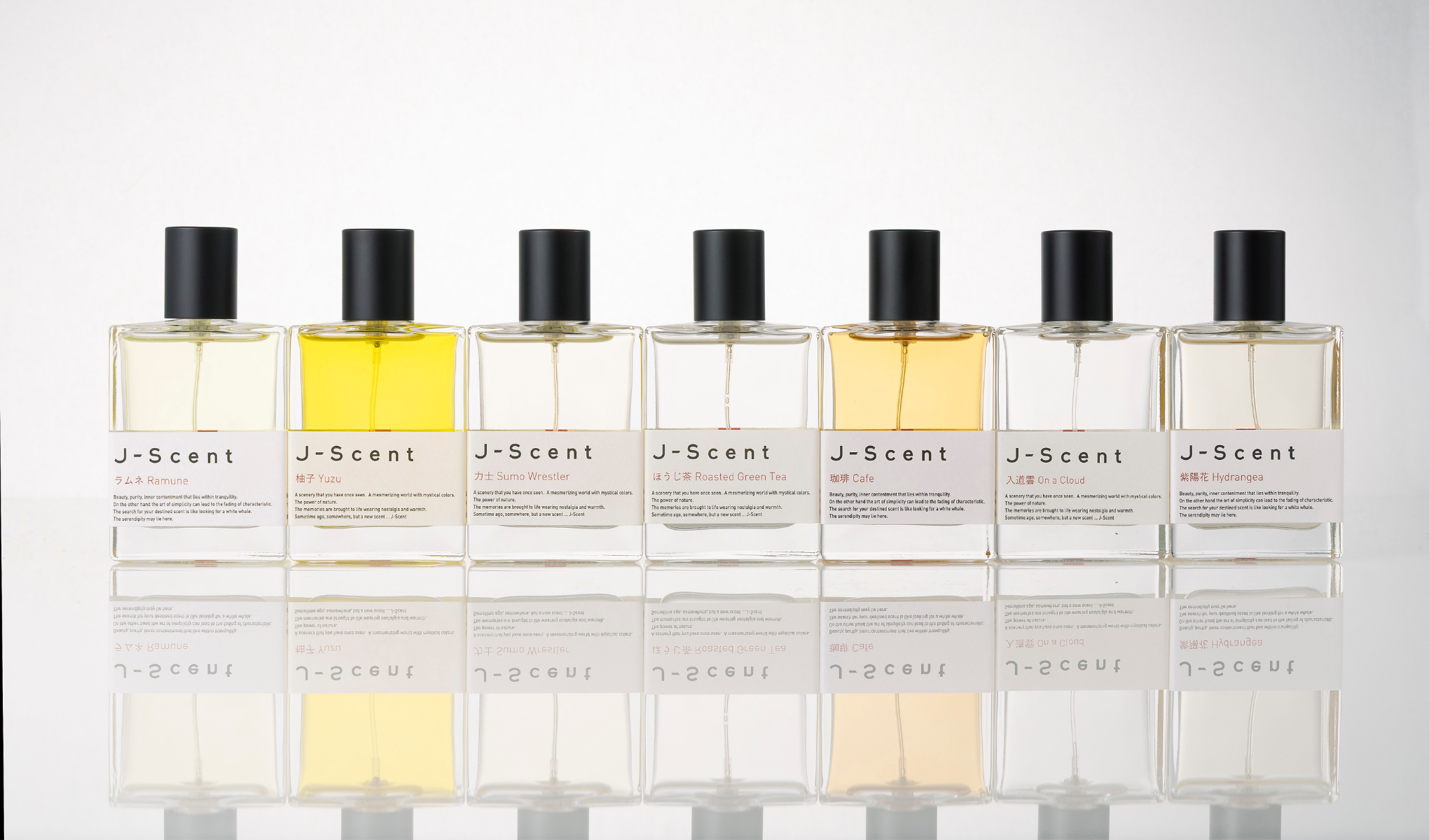 Tuotekuva seitsemästä J-Scent-hajuvedestä lasipulloissa mustilla korkeilla vaaleaa taustaa ja peiliheijastusta vasten. Näkyvät tuotenimet ovat “Ramune”, “Yuzu”, “Sumo Wrestler”, “Roasted Green Tea”, “Cafe”, “On a Cloud” ja “Hydrangea”.