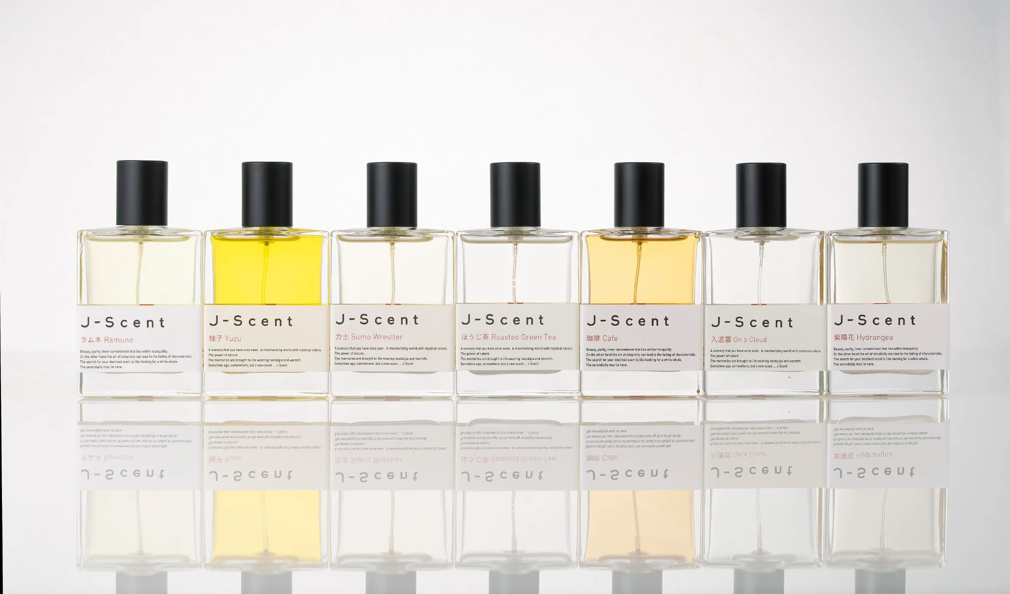 Produktbild med sju J-Scent-parfymer i glasflaskor med svarta lock mot ljus bakgrund med spegeleffekt. Synliga produktnamn inkluderar “Ramune”, “Yuzu”, “Sumo Wrestler”, “Roasted Green Tea”, “Cafe”, “On a Cloud” och “Hydrangea”.
