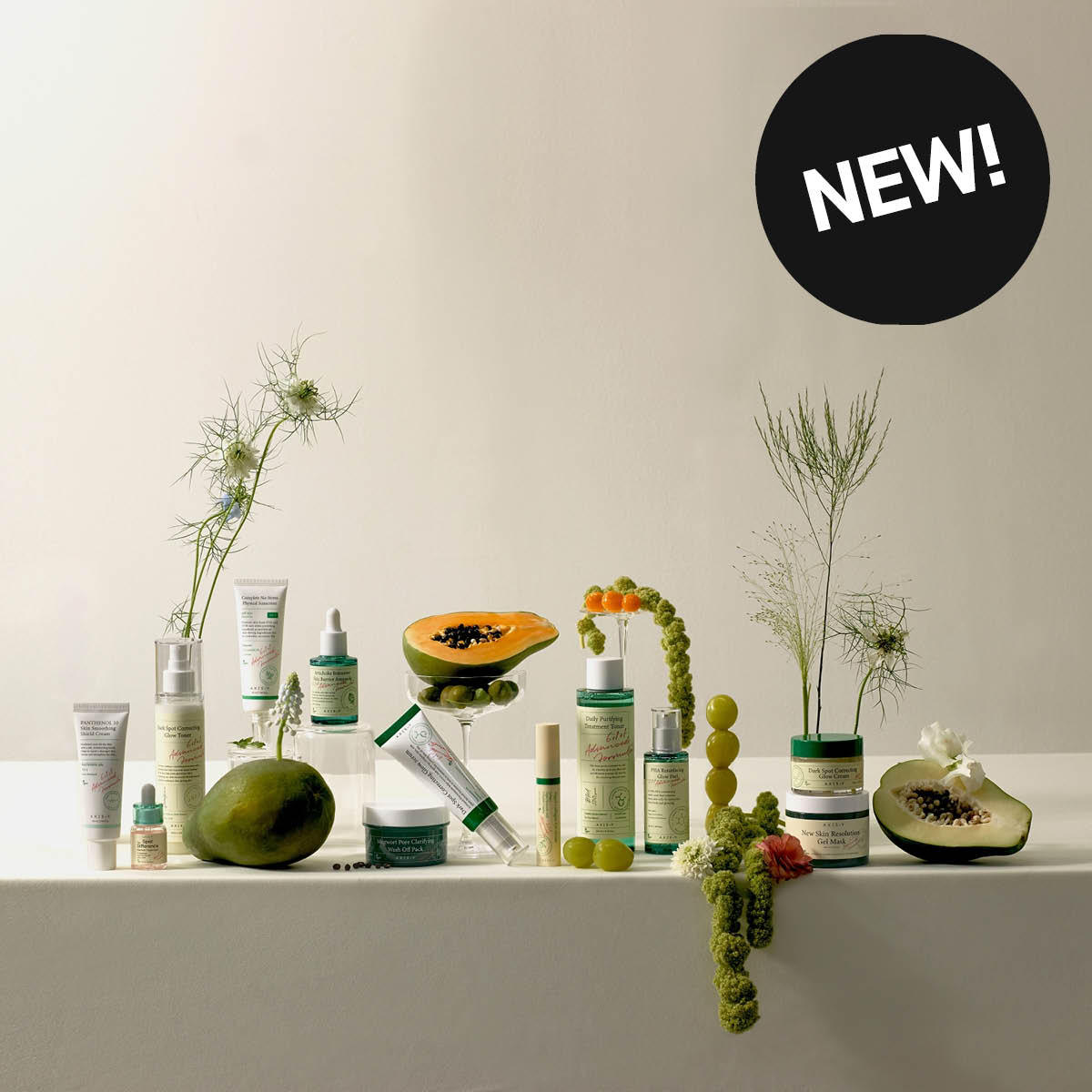 Et stort oppsett med hudpleieprodukter fra AXIS-Y, blant annet “Dark Spot Correcting Glow Toner”, “Dark Spot Correcting Glow Serum”, “Dark Spot Correcting Glow Cream” og “New Skin Resolution Gel Mask”, plassert blant frukt, glass og planter. En svart sirkel med teksten “NEW!” vises.