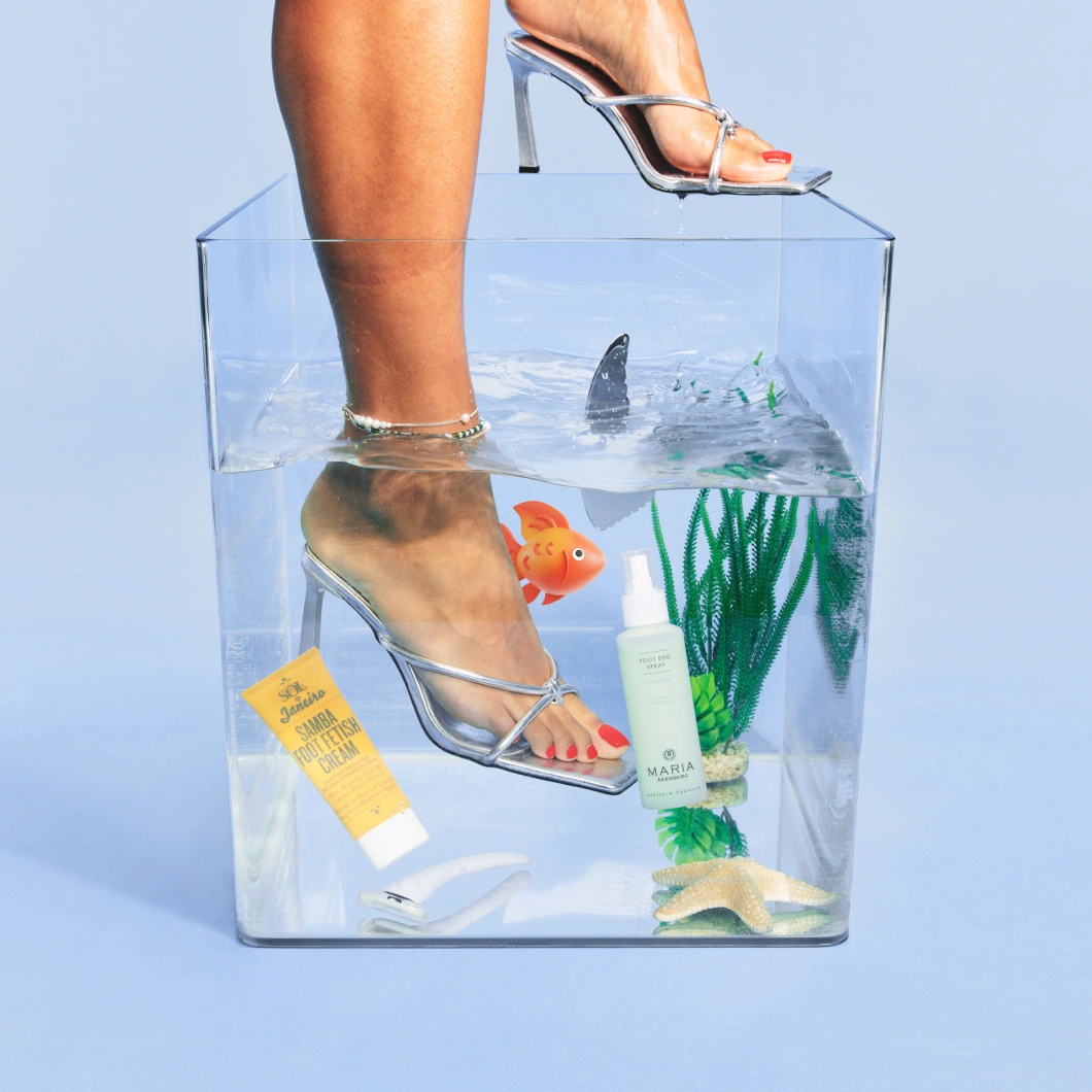 Person med sølvfargede høyhælte sandaler står i en gjennomsiktig vannfylt boks sammen med fotpleieprodukter, plastfisk, sjøstjerne og vannplanter. En gul tube med teksten “Sandal Foot Fetish Cream” og en lysegrønn sprayflaske med “Foot Deo Spray” fra Maria Åkerberg vises i bildet.