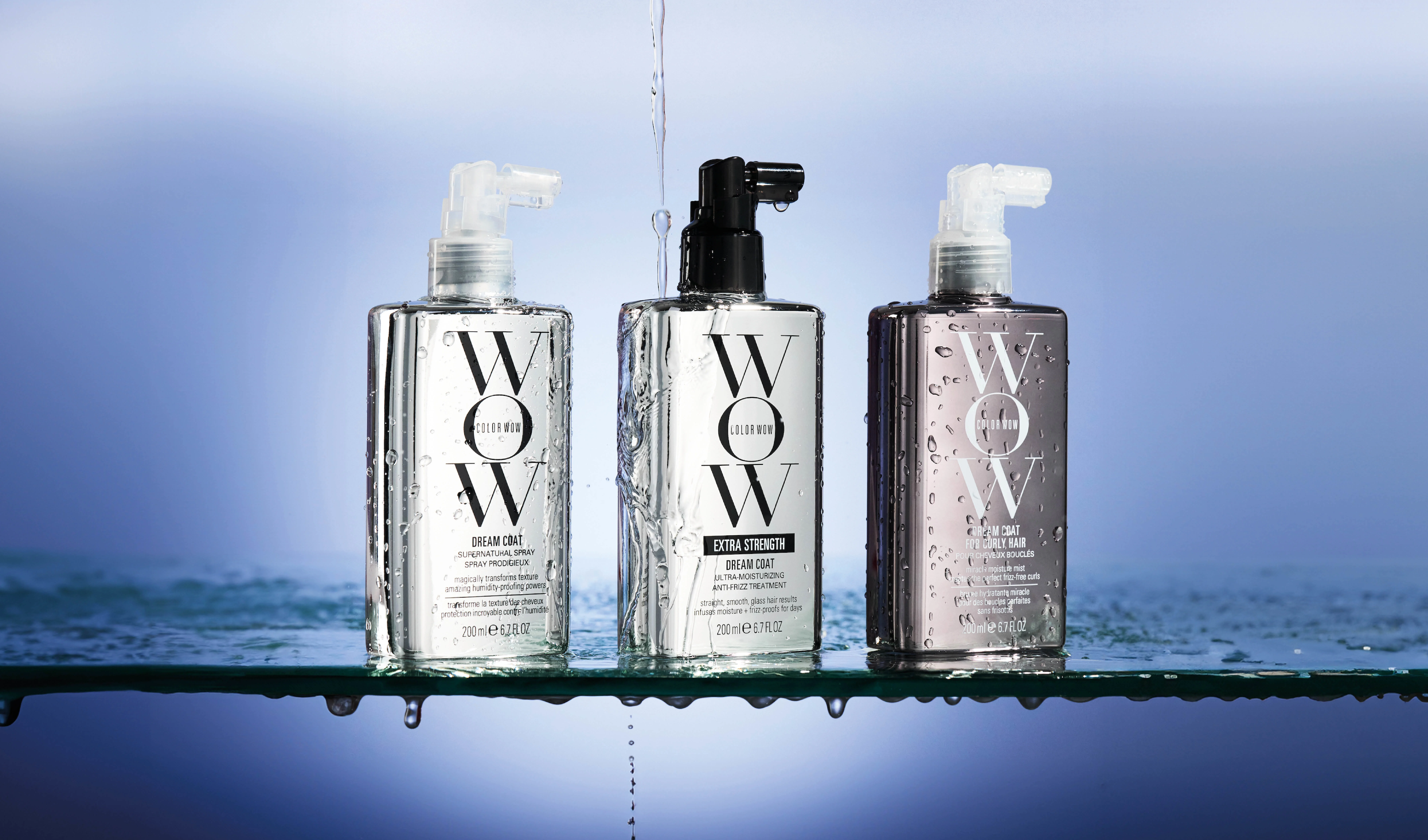 Tre flasker med Color WOW-hårprodukter, plassert på en våt overflate med vanndråper. Produktene er «Dream Coat Supernatural Spray», «Dream Coat Extra Strength» og «Dream Coat for Curly Hair». Bakgrunnen er blå, og flaskene er gjennomsiktige med etiketter i ulike farger.