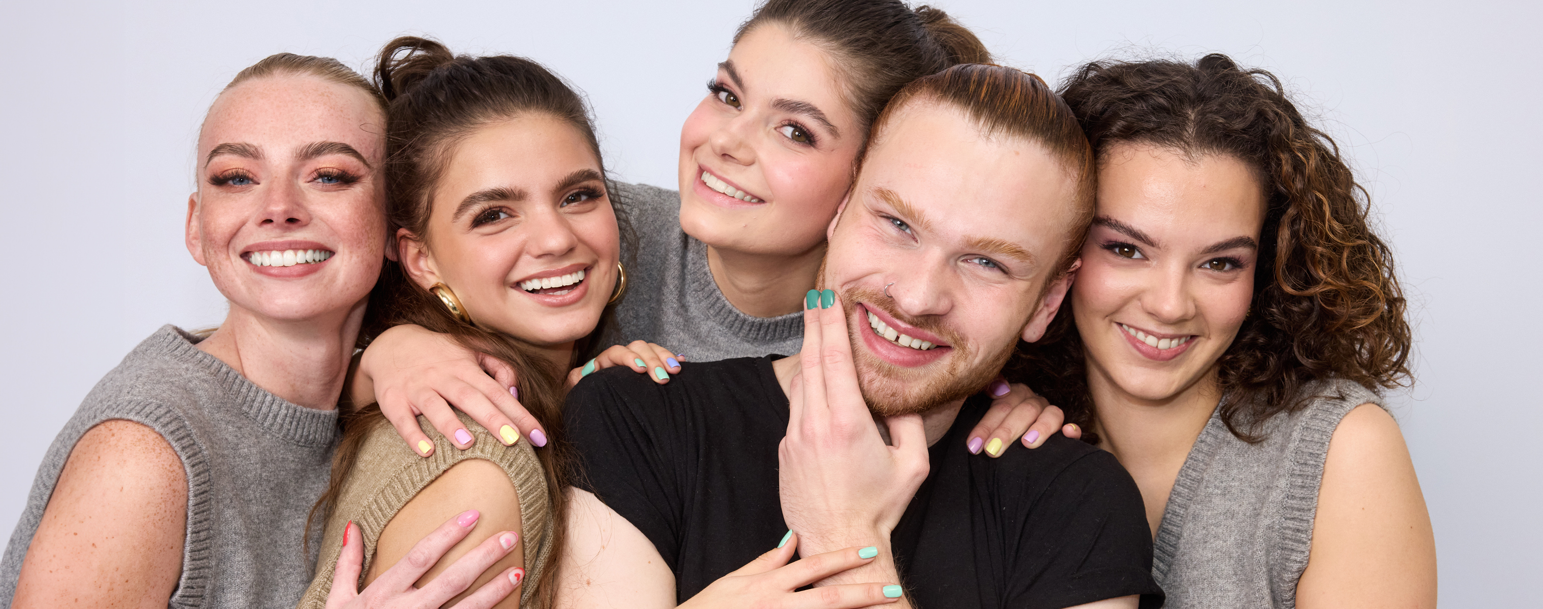 Fem personer står tett sammen og smiler. De har armene rundt hverandre og viser frem de fargerike neglene sine. Bakgrunnen er lys og nøytral.