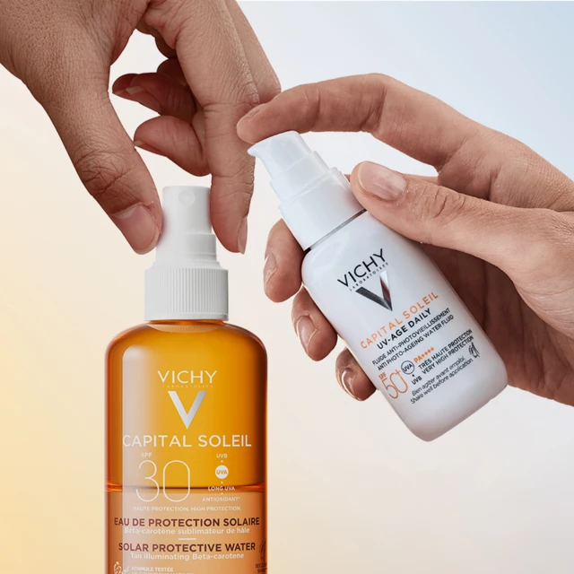 Två händer som håller två solskyddprodukter från Vichy mot en gulblå vägg.